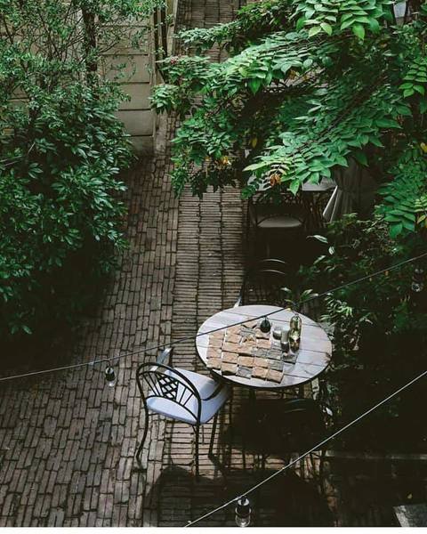 B&agrave;n tr&ograve;n với ghế đặt ngo&agrave;i trời tr&ecirc;n lối đi l&aacute;t gạch, xung quanh l&agrave; c&acirc;y xanh tươi tốt.