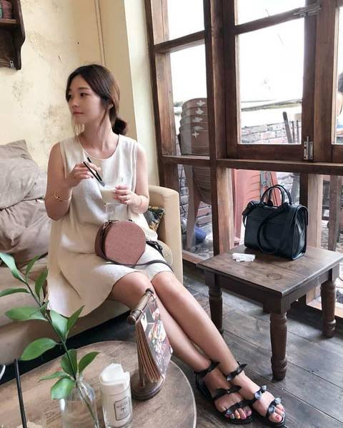 Người phụ nữ ngồi trong qu&aacute;n c&agrave; ph&ecirc;, cầm k&iacute;nh, b&ecirc;n cạnh t&uacute;i x&aacute;ch v&agrave; b&agrave;n gỗ, kh&ocirc;ng gian ấm &aacute;p.