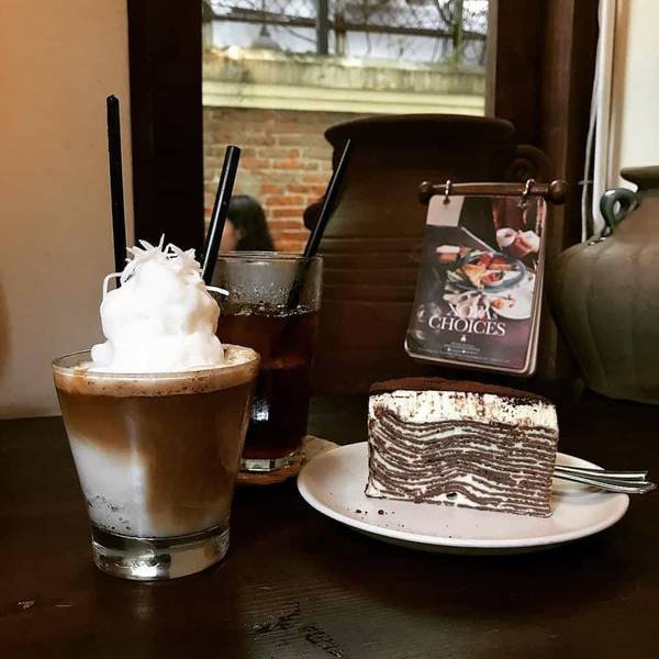 Một ly c&agrave; ph&ecirc;, một ly nước c&oacute; đ&aacute; v&agrave; một miếng b&aacute;nh nhiều lớp tr&ecirc;n b&agrave;n gỗ, gần cửa sổ.