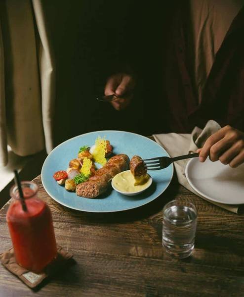 Người đang d&ugrave;ng dao nĩa ăn m&oacute;n ăn tr&ecirc;n đĩa m&agrave;u xanh, b&ecirc;n cạnh l&agrave; ly nước &eacute;p đỏ v&agrave; ly nước.