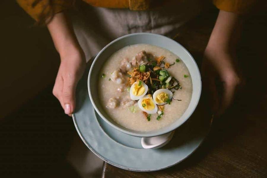 Một t&ocirc; ch&aacute;o n&oacute;ng hổi, trang tr&iacute; với trứng c&uacute;t luộc, h&agrave;nh l&aacute; v&agrave; h&agrave;nh phi, được bưng bởi hai b&agrave;n tay.