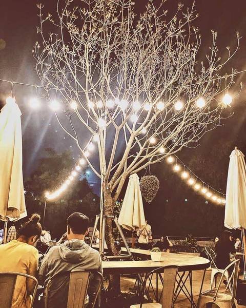 Qu&aacute;n c&agrave; ph&ecirc; ngo&agrave;i trời v&agrave;o buổi tối, c&oacute; c&acirc;y trang tr&iacute; đ&egrave;n, b&agrave;n ghế kim loại v&agrave; một số người ngồi thư gi&atilde;n.