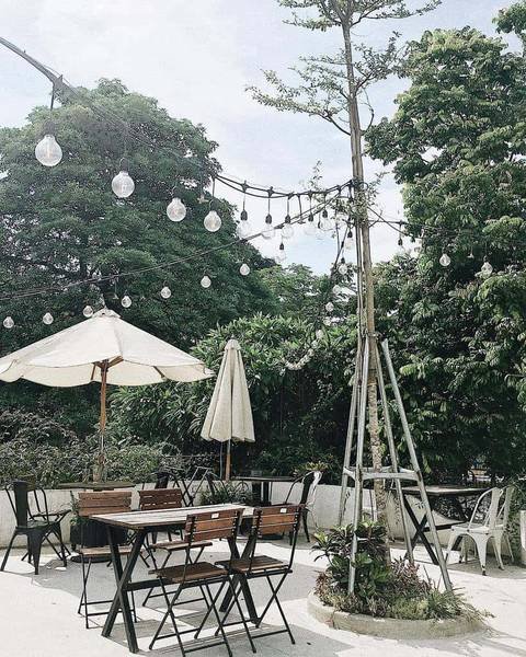 Qu&aacute;n c&agrave; ph&ecirc; ngo&agrave;i trời với c&acirc;y xanh, b&agrave;n ghế gỗ, d&ugrave; che trắng v&agrave; d&acirc;y đ&egrave;n treo trang tr&iacute; tạo kh&ocirc;ng gian tho&aacute;ng đ&atilde;ng.