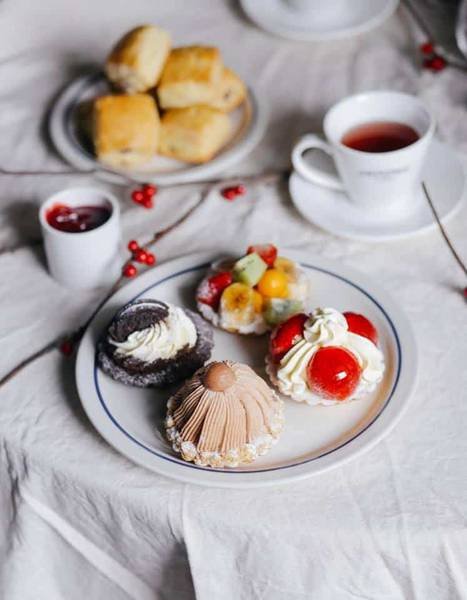 Ba chiếc b&aacute;nh ngọt tr&ecirc;n đĩa, k&egrave;m t&aacute;ch tr&agrave; v&agrave; b&aacute;nh nướng tr&ecirc;n b&agrave;n trải khăn trắng.