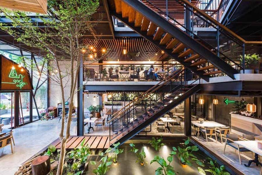 Kh&ocirc;ng gian qu&aacute;n c&agrave; ph&ecirc; hiện đại với cầu thang gỗ, c&acirc;y xanh v&agrave; &aacute;nh s&aacute;ng tự nhi&ecirc;n từ cửa sổ lớn.
