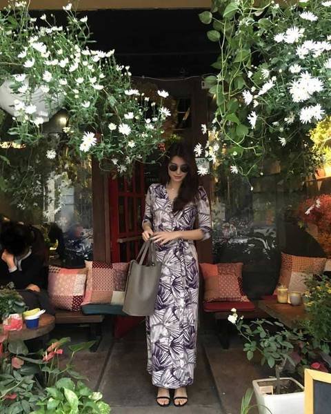 Người phụ nữ mặc v&aacute;y d&agrave;i, đeo k&iacute;nh r&acirc;m, đứng trước qu&aacute;n c&agrave; ph&ecirc; với nhiều c&acirc;y xanh v&agrave; hoa treo.