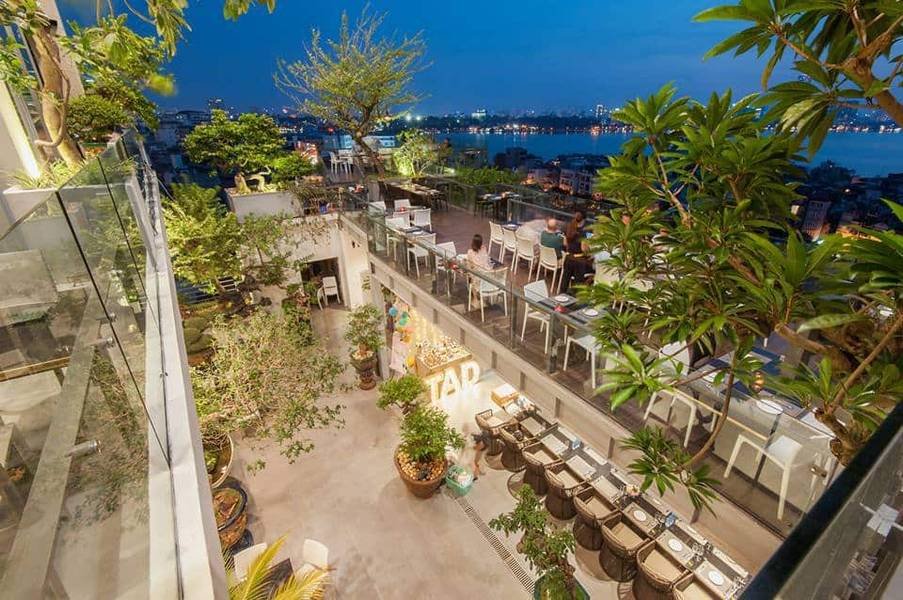 Qu&aacute;n c&agrave; ph&ecirc; ngo&agrave;i trời tr&ecirc;n tầng cao với b&agrave;n ghế, c&acirc;y xanh, nh&igrave;n ra hồ v&agrave; th&agrave;nh phố v&agrave;o buổi tối.