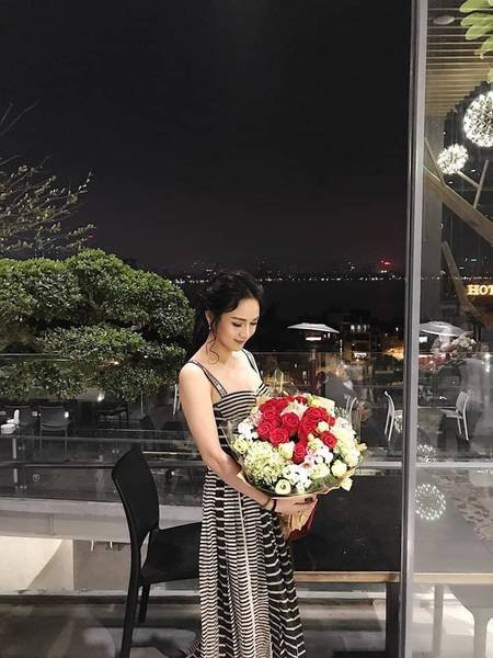 Người phụ nữ mặc v&aacute;y kẻ &ocirc;m b&oacute; hoa lớn, đứng ngo&agrave;i trời v&agrave;o buổi tối, ph&iacute;a sau l&agrave; cảnh th&agrave;nh phố lung linh &aacute;nh đ&egrave;n.