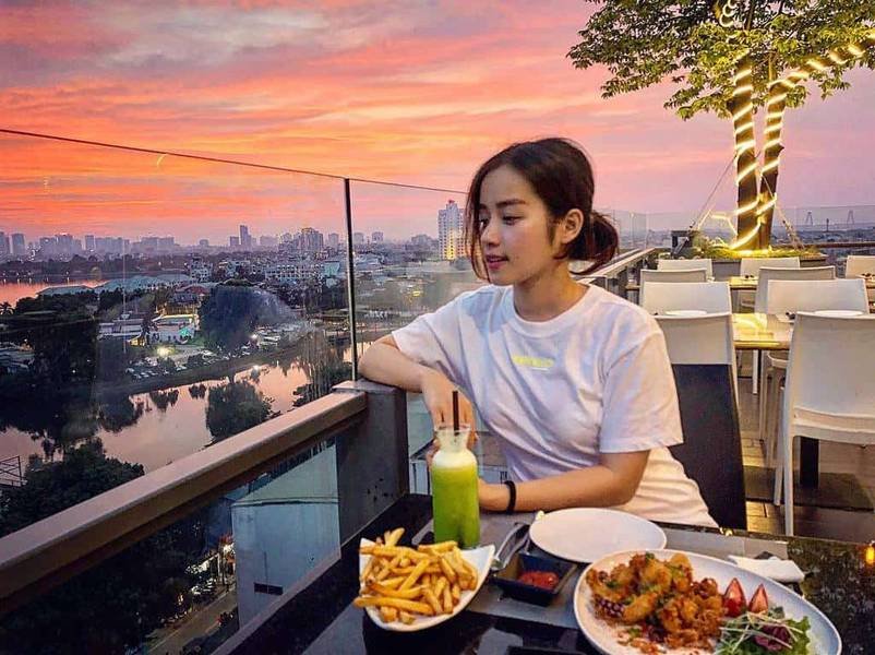 Người ngồi tr&ecirc;n ban c&ocirc;ng qu&aacute;n c&agrave; ph&ecirc;, trước mặt l&agrave; đồ ăn v&agrave; nước uống, ph&iacute;a sau l&agrave; cảnh ho&agrave;ng h&ocirc;n th&agrave;nh phố.