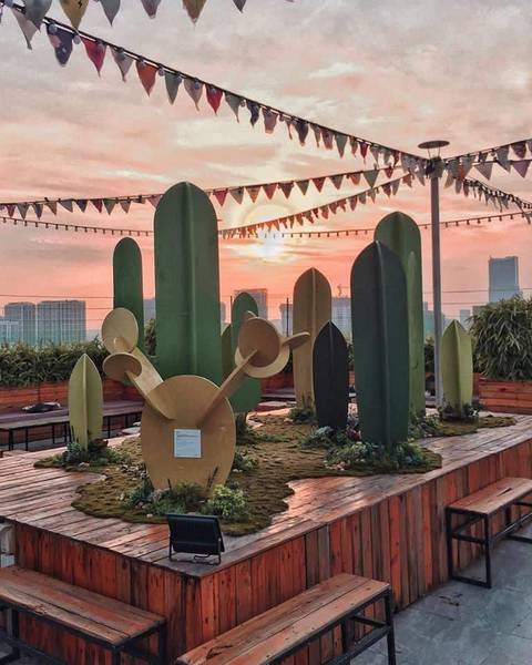 Khu vườn s&acirc;n thượng với m&ocirc; h&igrave;nh xương rồng v&agrave; b&agrave;n ghế gỗ, ph&ocirc;ng nền l&agrave; ho&agrave;ng h&ocirc;n v&agrave; t&ograve;a nh&agrave; cao tầng.