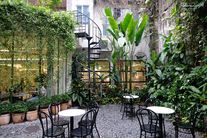 Quán cà phê ngoài trời với bàn ghế, cây xanh và cầu thang xoắn, tạo không gian thư giãn và thoáng đãng.