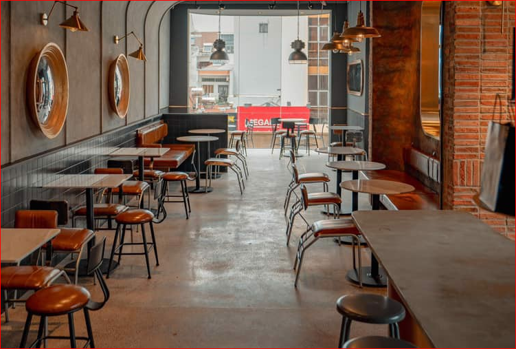 Quán cà phê với bàn ghế xếp gọn, ánh sáng tự nhiên từ cửa sổ lớn, trang trí đèn treo và gương tròn.