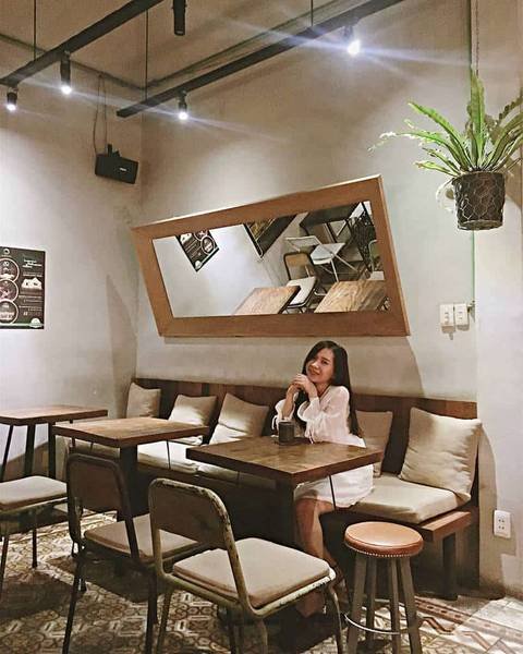 Qu&aacute;n c&agrave; ph&ecirc; với b&agrave;n ghế gỗ, đ&egrave;n treo v&agrave; gương lớn nghi&ecirc;ng, c&oacute; một người đang ngồi thư gi&atilde;n.