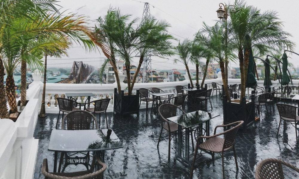 Qu&aacute;n c&agrave; ph&ecirc; ngo&agrave;i trời với b&agrave;n ghế v&agrave; c&acirc;y cọ, trời mưa, nh&igrave;n ra cảnh th&agrave;nh phố v&agrave; c&acirc;y cầu xa xa.