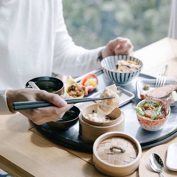 Người ngồi ăn bữa ăn kiểu &Aacute; với c&aacute;c m&oacute;n ăn đa dạng tr&ecirc;n b&agrave;n, d&ugrave;ng đũa gắp b&aacute;nh bao từ xửng tre.