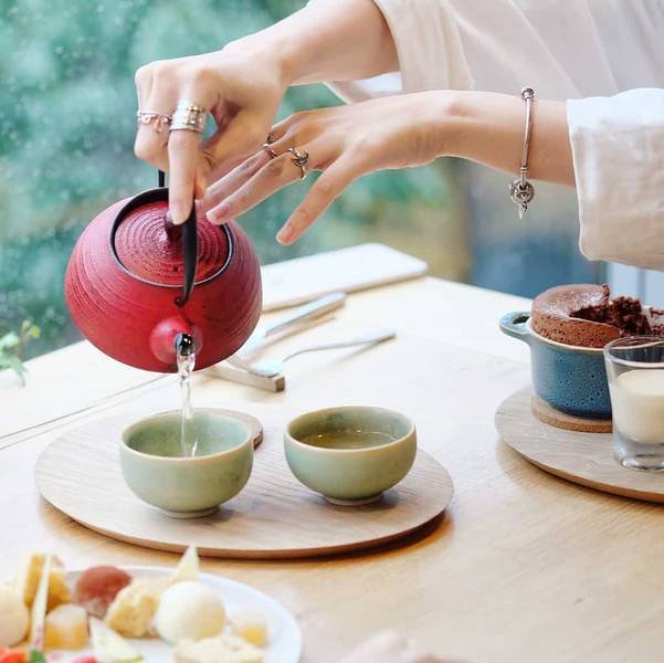 Một người đang r&oacute;t tr&agrave; từ ấm đỏ v&agrave;o t&aacute;ch tr&ecirc;n b&agrave;n gỗ, cạnh đ&oacute; l&agrave; đồ ăn v&agrave; đồ uống kh&aacute;c.