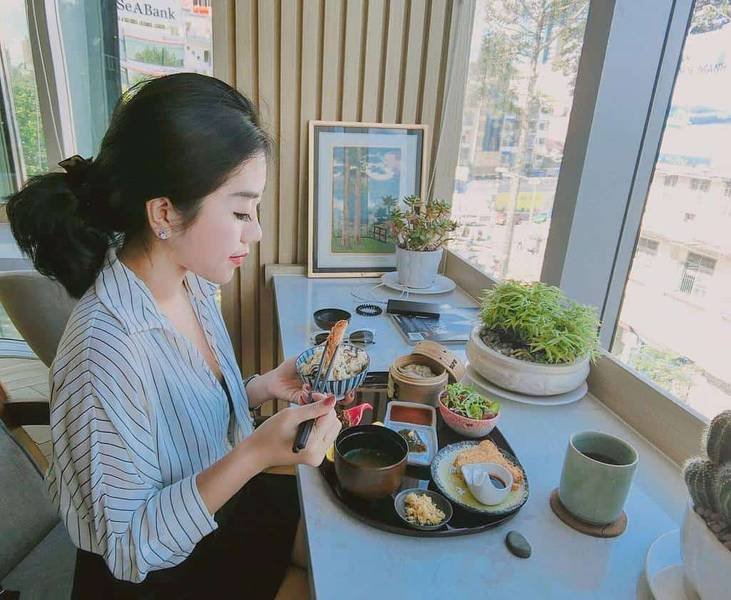 Người phụ nữ ngồi ăn trưa tại qu&aacute;n c&agrave; ph&ecirc;, b&ecirc;n cửa sổ c&oacute; c&acirc;y xanh v&agrave; nh&igrave;n ra khung cảnh đường phố.