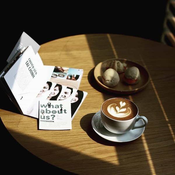 T&aacute;ch c&agrave; ph&ecirc; latte v&agrave; đĩa b&aacute;nh ngọt tr&ecirc;n b&agrave;n gỗ, b&ecirc;n cạnh l&agrave; s&aacute;ch v&agrave; tạp ch&iacute; mở dưới &aacute;nh s&aacute;ng tự nhi&ecirc;n.