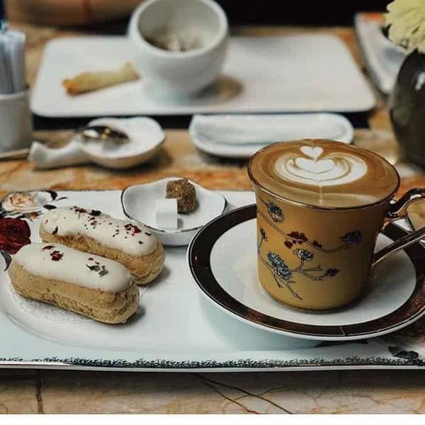 T&aacute;ch c&agrave; ph&ecirc; latte v&agrave; b&aacute;nh ngọt tr&ecirc;n đĩa trắng, trang tr&iacute; tinh tế với hoa văn.