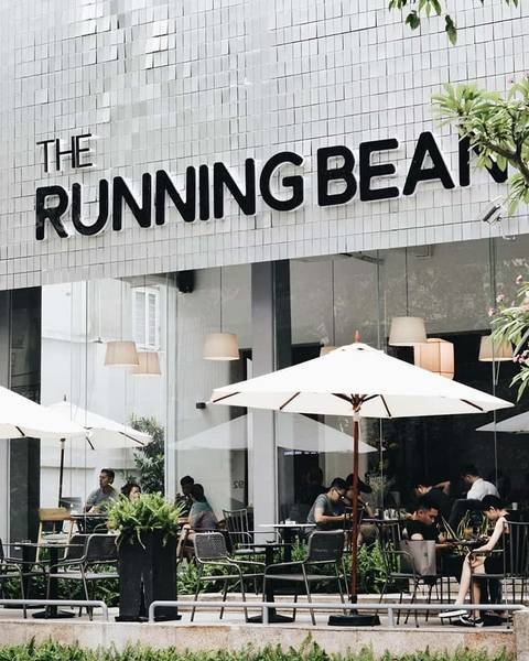 Qu&aacute;n c&agrave; ph&ecirc; với bảng hiệu "The Running Bean", c&oacute; kh&aacute;ch ngồi ngo&agrave;i trời dưới d&ugrave; trắng.