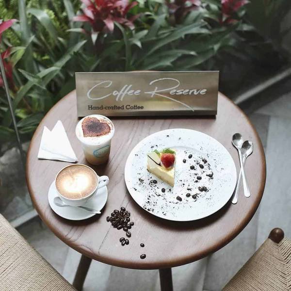 B&agrave;n tr&ograve;n với c&agrave; ph&ecirc;, b&aacute;nh kem, v&agrave; tấm bảng "Coffee Reserve" tr&ecirc;n nền hoa l&aacute; xanh.