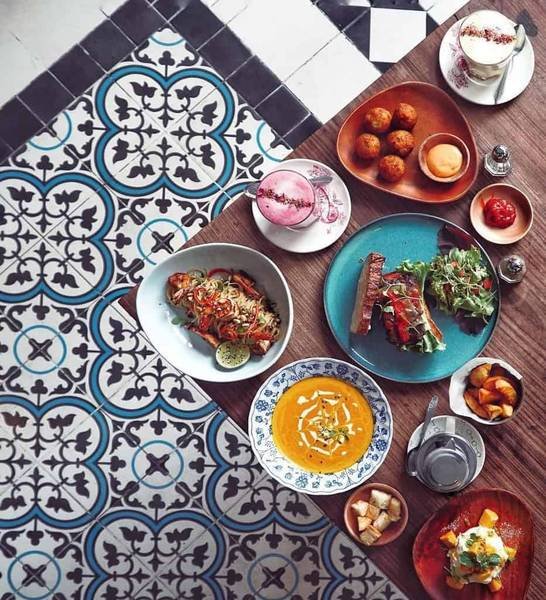 B&agrave;n ăn với nhiều m&oacute;n ăn đa dạng tr&ecirc;n đĩa m&agrave;u sắc, đặt tr&ecirc;n nền gạch hoa văn trang tr&iacute; tinh tế.