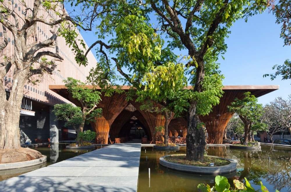 Lối đi dẫn v&agrave;o t&ograve;a nh&agrave; c&oacute; kiến tr&uacute;c tre, xung quanh l&agrave; c&acirc;y xanh v&agrave; hồ nước, tạo kh&ocirc;ng gian xanh m&aacute;t.