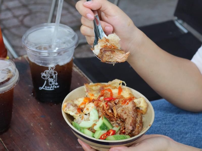 Một người đang cầm b&aacute;t cơm tấm với thịt, dưa leo v&agrave; ớt, b&ecirc;n cạnh l&agrave; hai ly nước uống tr&ecirc;n b&agrave;n.