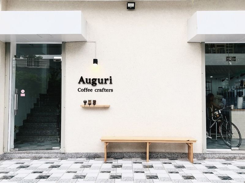 Mặt tiền qu&aacute;n c&agrave; ph&ecirc; với biển hiệu "Auguri Coffee Crafters", cửa k&iacute;nh v&agrave; ghế gỗ d&agrave;i ph&iacute;a trước.