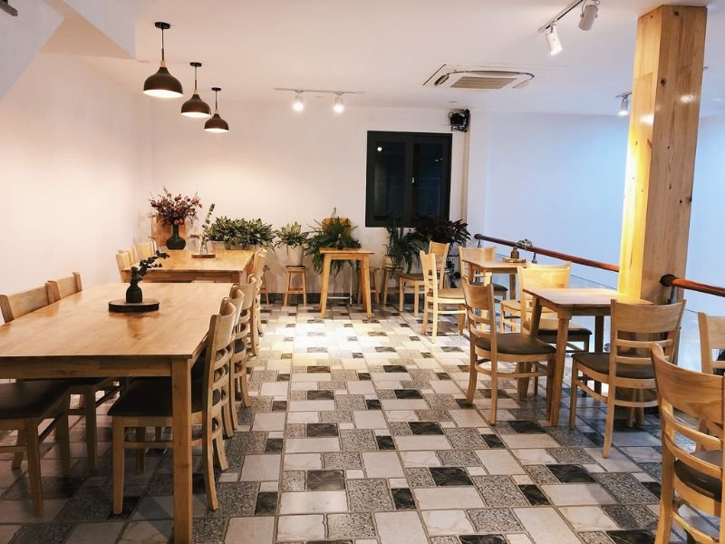 Qu&aacute;n c&agrave; ph&ecirc; với b&agrave;n ghế gỗ, s&agrave;n l&aacute;t gạch, trang tr&iacute; c&acirc;y xanh v&agrave; đ&egrave;n treo trần, tạo kh&ocirc;ng gian ấm c&uacute;ng.