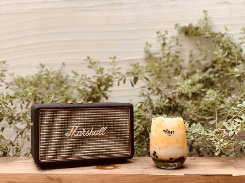 Loa di động v&agrave; ly tr&agrave; sữa tr&acirc;n ch&acirc;u tr&ecirc;n b&agrave;n gỗ, nền c&acirc;y xanh ph&iacute;a sau.