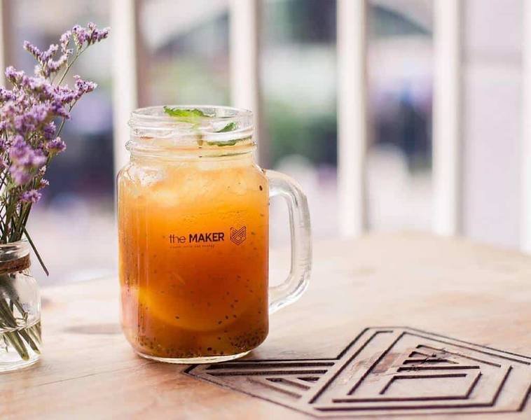 Ly nước cam trong suốt trên bàn gỗ, bên cạnh là lọ hoa tím nhỏ, tạo cảm giác thư giãn.