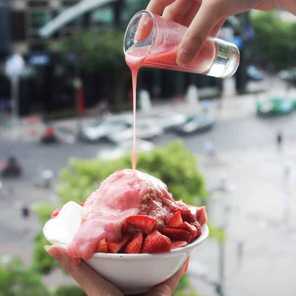 Người đang rót nước sốt dâu tây lên bát đá bào với dâu tươi, khung cảnh ngoài trời phía sau.