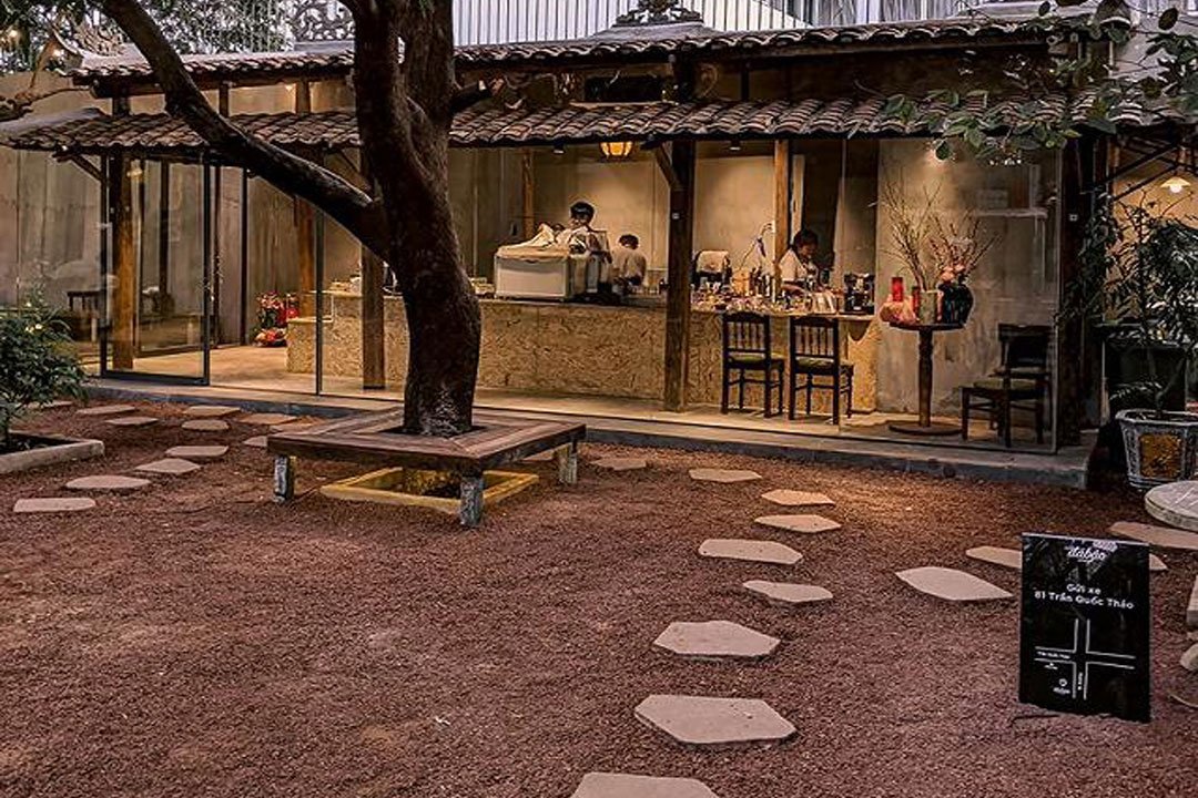 Quán cà phê với mái ngói cổ điển, có lối đi đá, cây lớn giữa sân, nội thất gỗ và cửa kính.