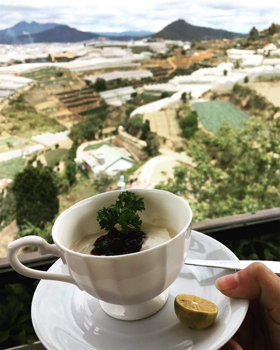 T&aacute;ch tr&agrave; với m&oacute;n tr&aacute;ng miệng v&agrave; miếng chanh, nền l&agrave; khung cảnh thung lũng v&agrave; n&uacute;i non mờ ảo.