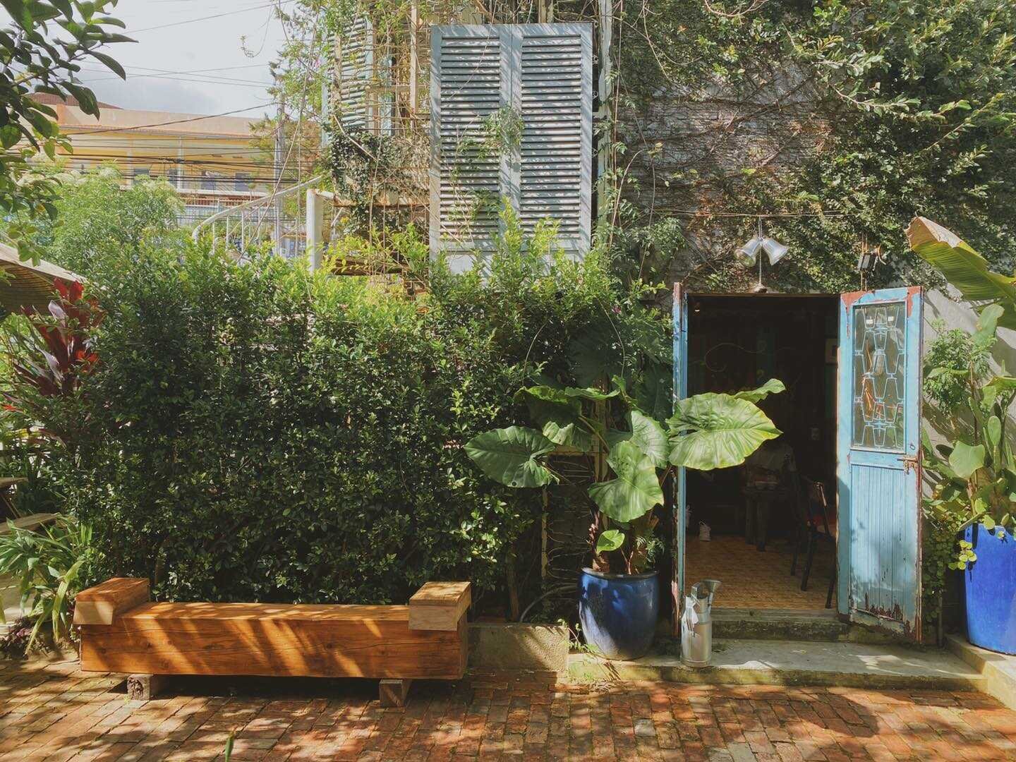 Khu vườn xanh tươi, cửa gỗ m&agrave;u xanh mở ra, ghế gỗ đặt tr&ecirc;n nền gạch, c&acirc;y cối bao quanh.
