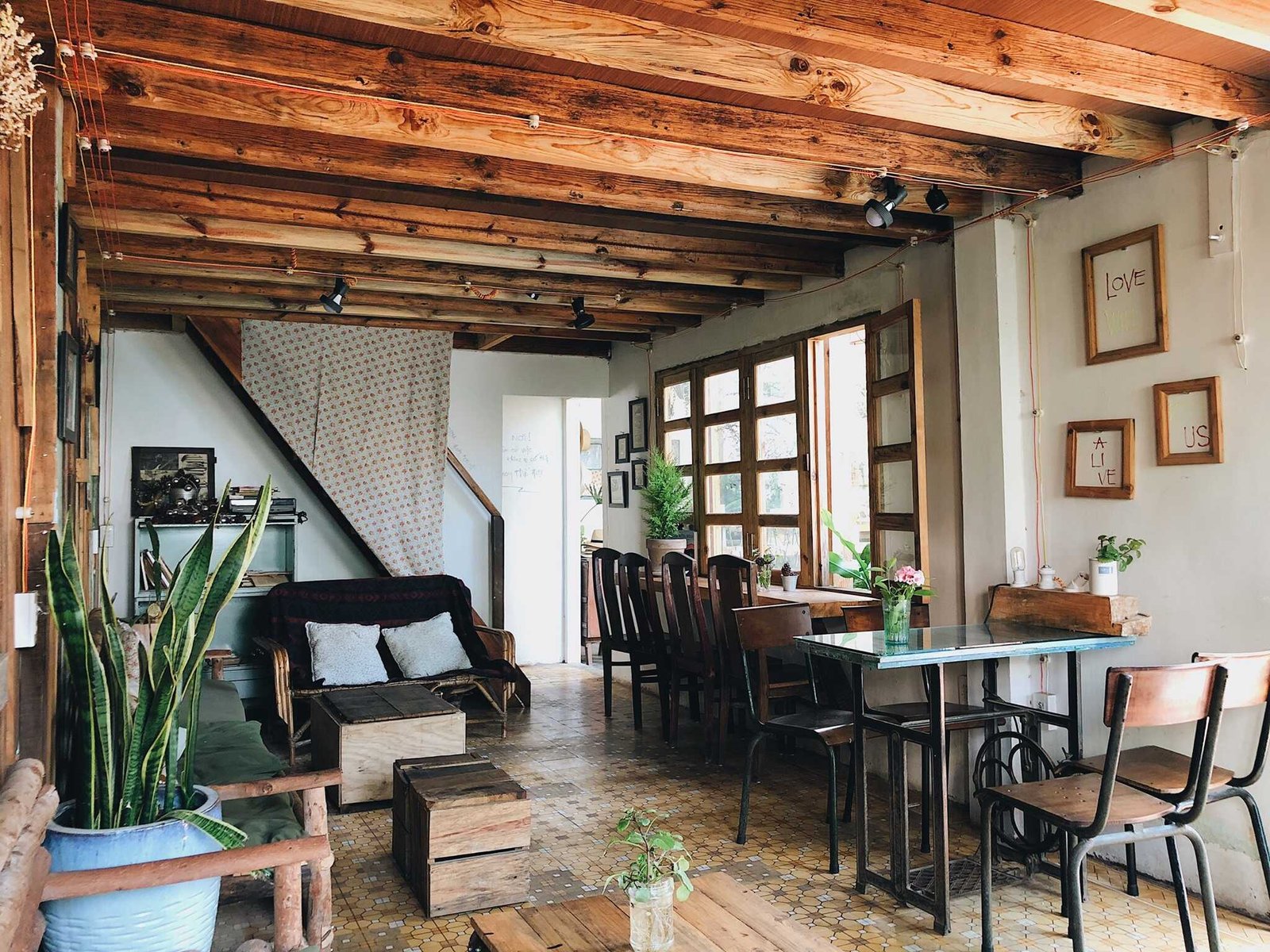 Qu&aacute;n c&agrave; ph&ecirc; với nội thất gỗ ấm c&uacute;ng, b&agrave;n ghế xếp gọn g&agrave;ng, c&acirc;y xanh v&agrave; tranh treo tr&ecirc;n tường.