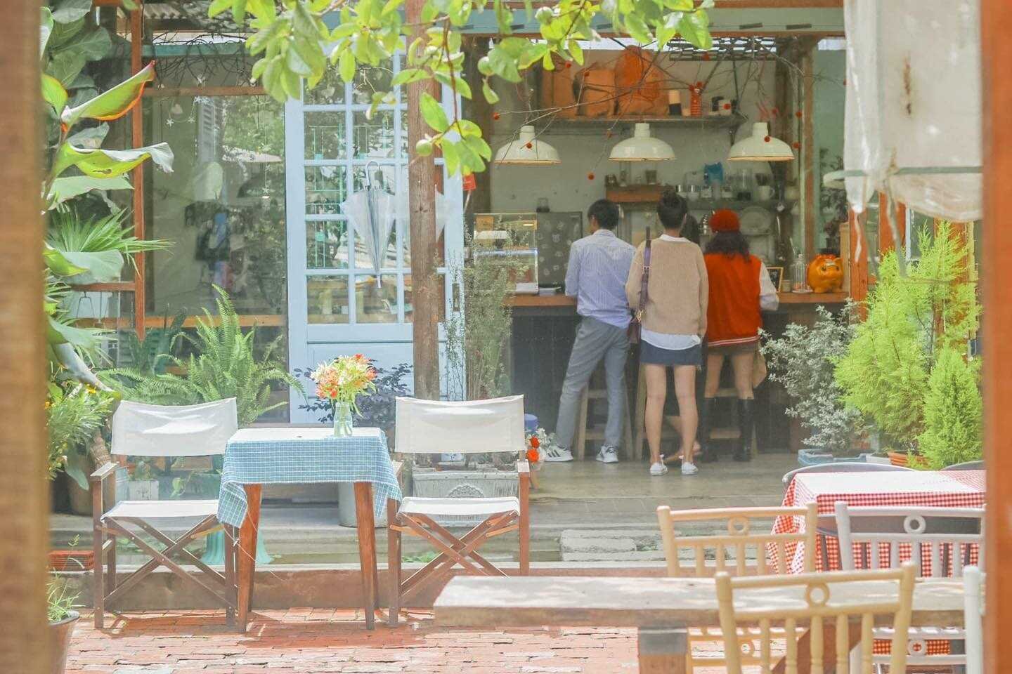 Qu&aacute;n c&agrave; ph&ecirc; ngo&agrave;i trời với b&agrave;n ghế gỗ, c&acirc;y xanh, v&agrave; ba người đứng b&ecirc;n quầy pha chế b&ecirc;n trong.