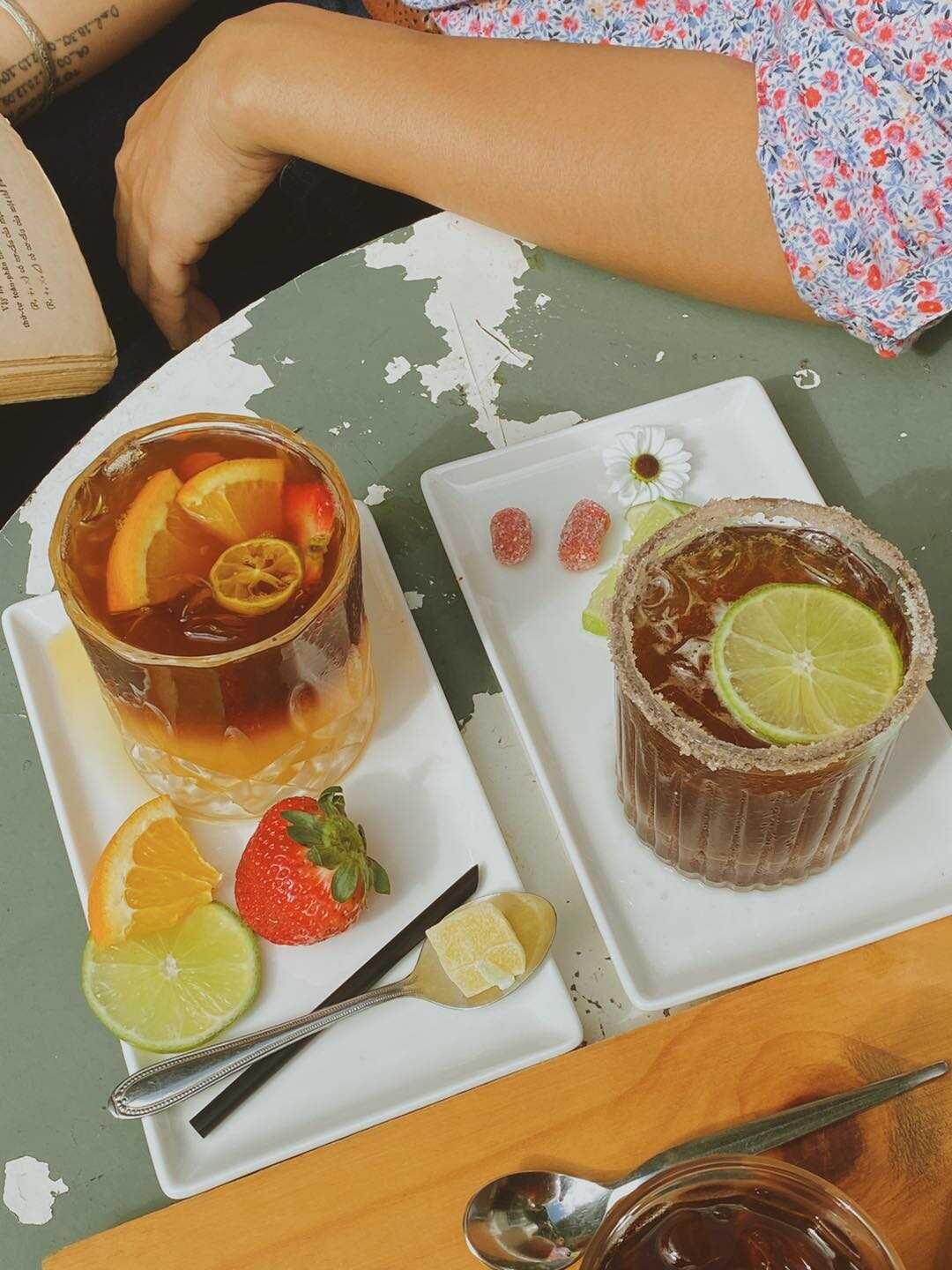 Hai ly nước uống trang tr&iacute; với l&aacute;t chanh, cam v&agrave; d&acirc;u t&acirc;y tr&ecirc;n b&agrave;n, b&ecirc;n cạnh l&agrave; một người cầm cuốn s&aacute;ch.