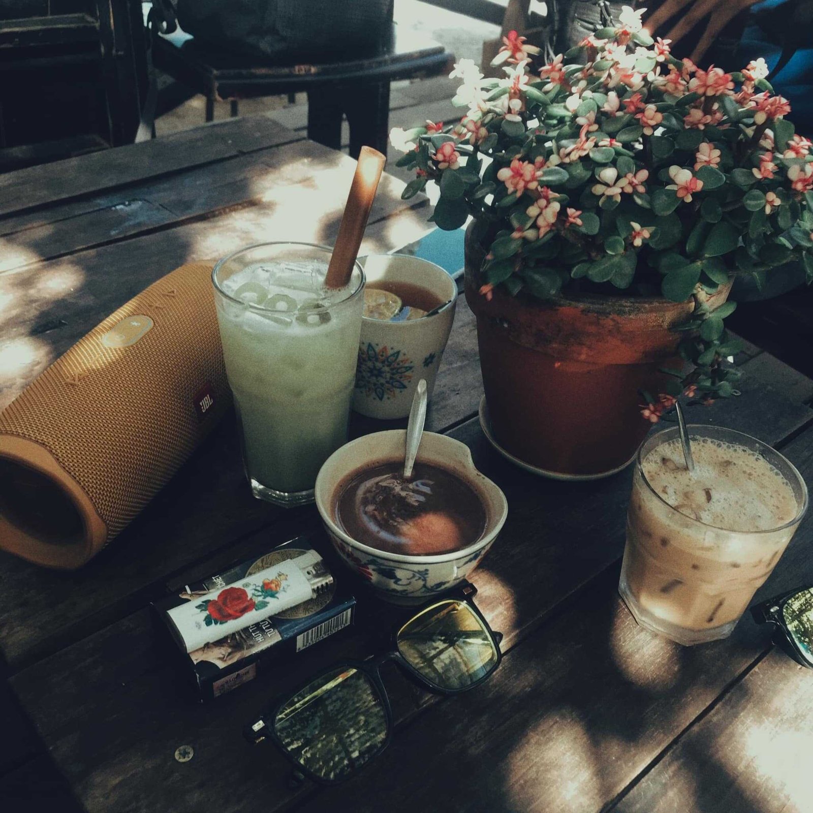 B&agrave;n gỗ c&oacute; c&acirc;y hoa, nhiều loại đồ uống v&agrave; vật dụng c&aacute; nh&acirc;n dưới &aacute;nh s&aacute;ng mặt trời.