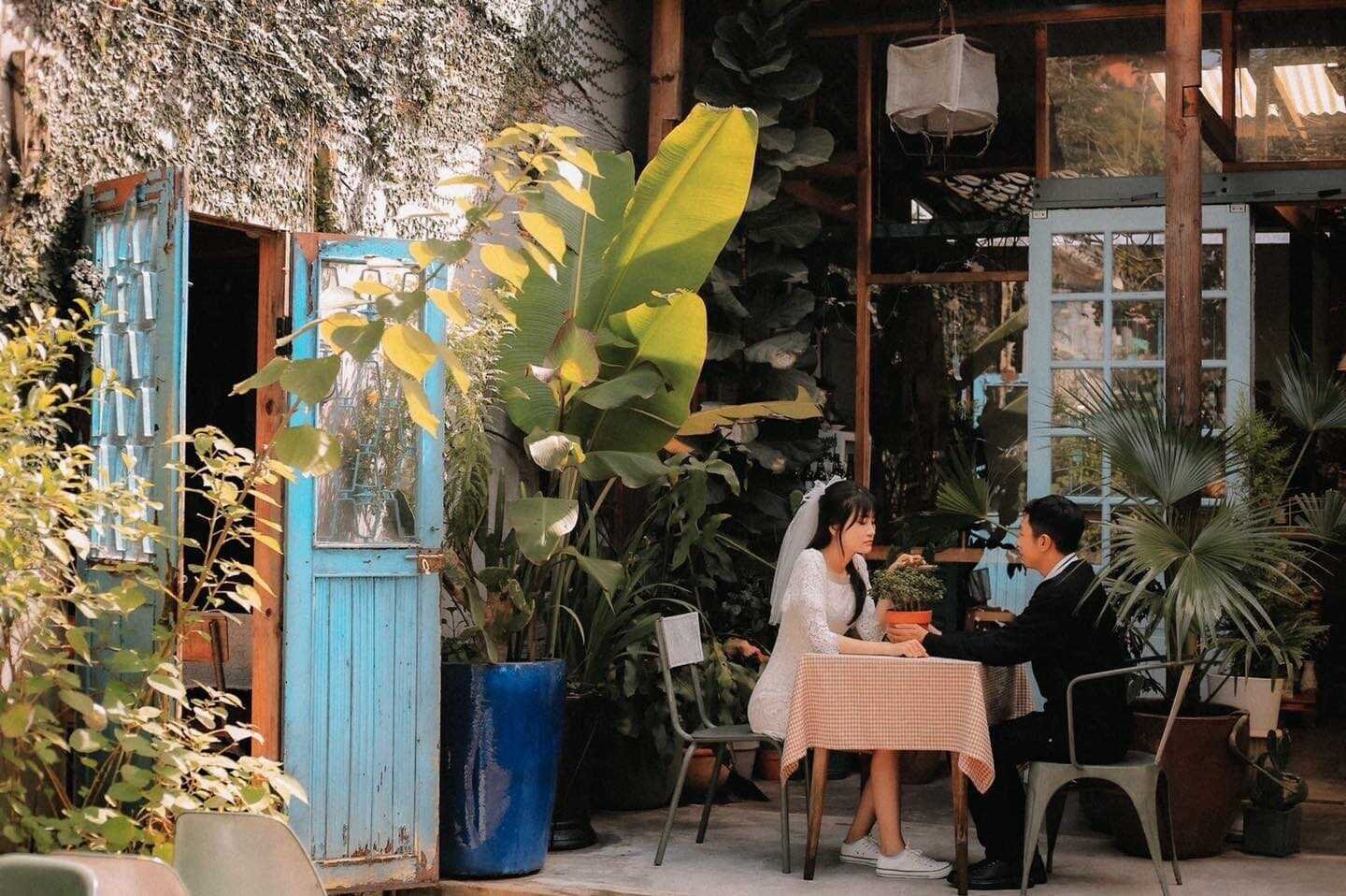 Cặp đ&ocirc;i ngồi ở b&agrave;n ngo&agrave;i trời, xung quanh l&agrave; c&acirc;y cối v&agrave; cửa gỗ m&agrave;u xanh.