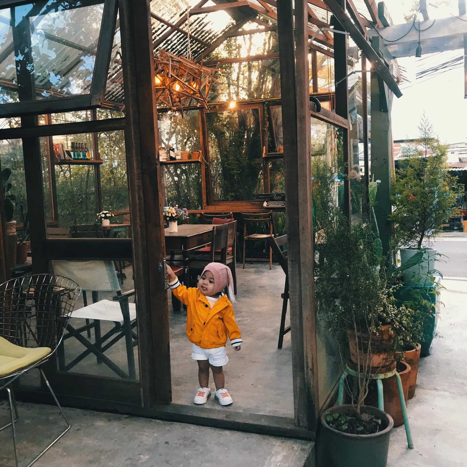 Em b&eacute; mặc &aacute;o v&agrave;ng đứng trước cửa k&iacute;nh của qu&aacute;n c&agrave; ph&ecirc;, xung quanh c&oacute; nhiều c&acirc;y xanh v&agrave; b&agrave;n ghế.