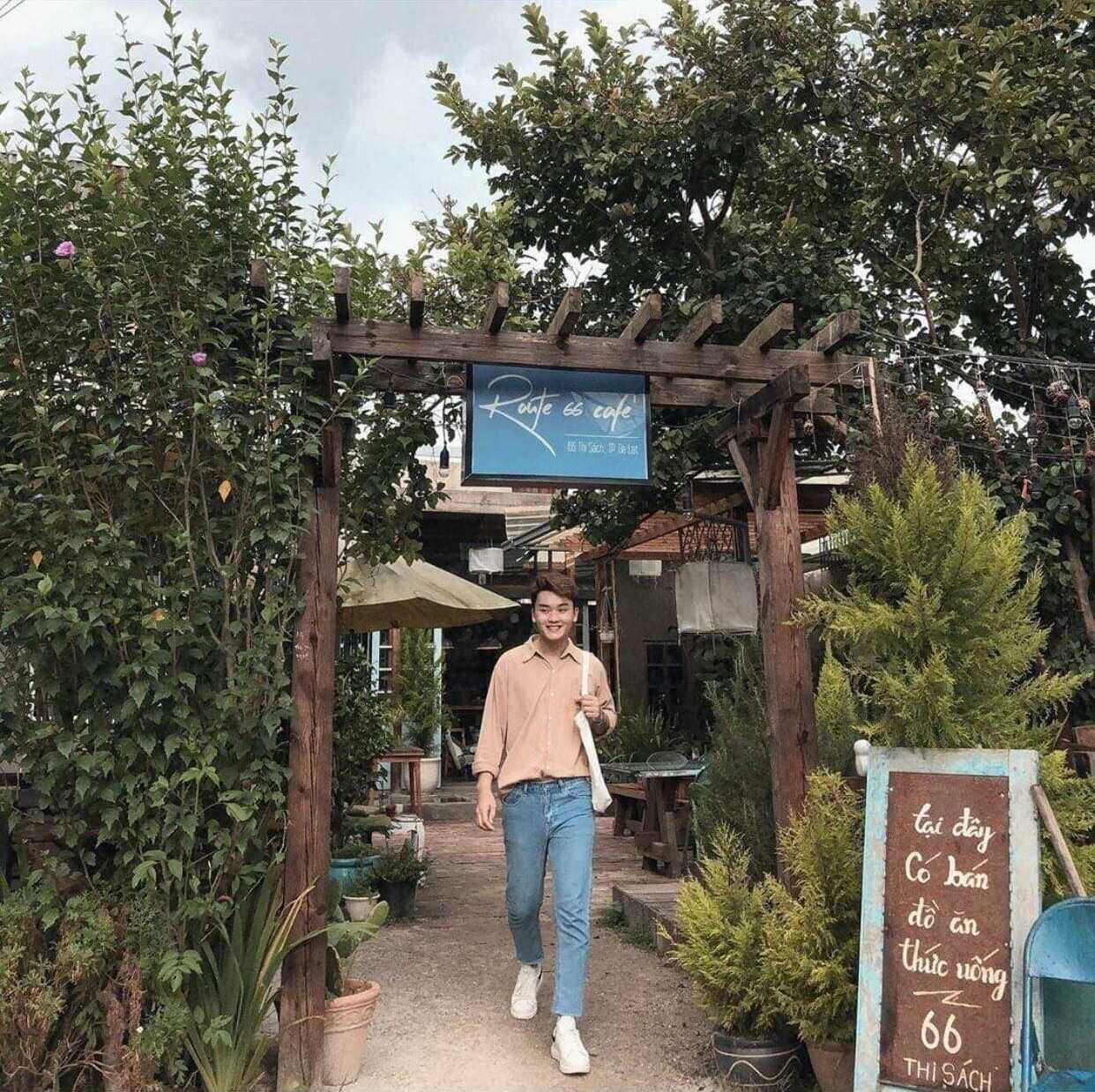 Người đi bộ dưới cổng gỗ của qu&aacute;n c&agrave; ph&ecirc; ngo&agrave;i trời, xung quanh c&oacute; nhiều c&acirc;y xanh v&agrave; bảng hiệu.