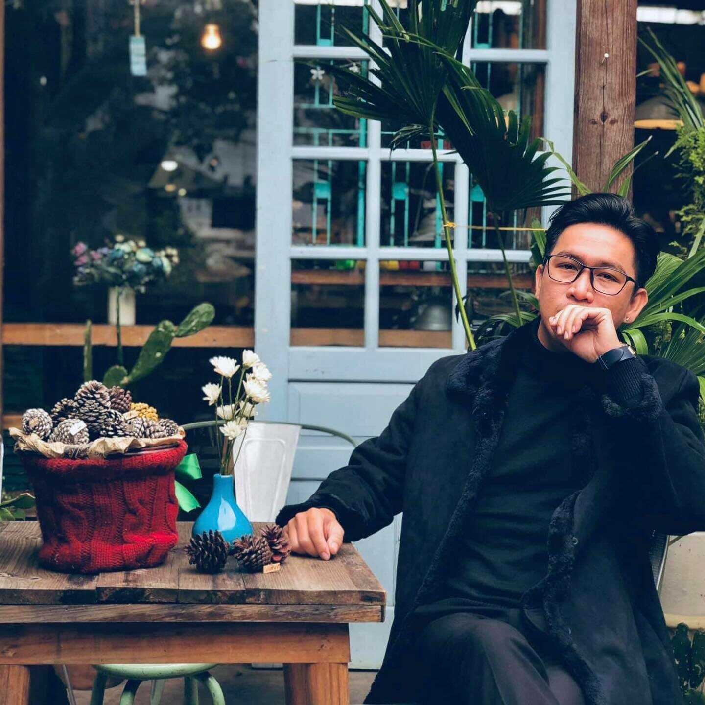 Người đ&agrave;n &ocirc;ng ngồi ngo&agrave;i trời b&ecirc;n b&agrave;n gỗ, cạnh chậu hoa v&agrave; quả th&ocirc;ng, nền l&agrave; cửa k&iacute;nh v&agrave; c&acirc;y xanh.