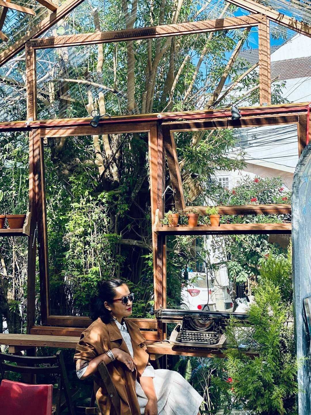 Người ngồi trong nh&agrave; k&iacute;nh với m&aacute;y đ&aacute;nh chữ cổ tr&ecirc;n b&agrave;n, xung quanh c&oacute; c&acirc;y xanh v&agrave; &aacute;nh s&aacute;ng tự nhi&ecirc;n.