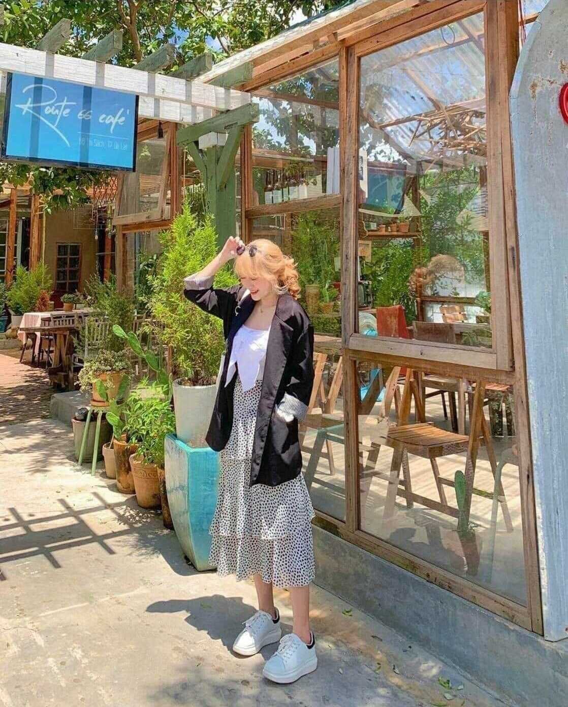 Người đứng trước qu&aacute;n c&agrave; ph&ecirc; ngo&agrave;i trời, mặc v&aacute;y chấm bi v&agrave; &aacute;o kho&aacute;c đen, xung quanh c&oacute; nhiều c&acirc;y xanh.