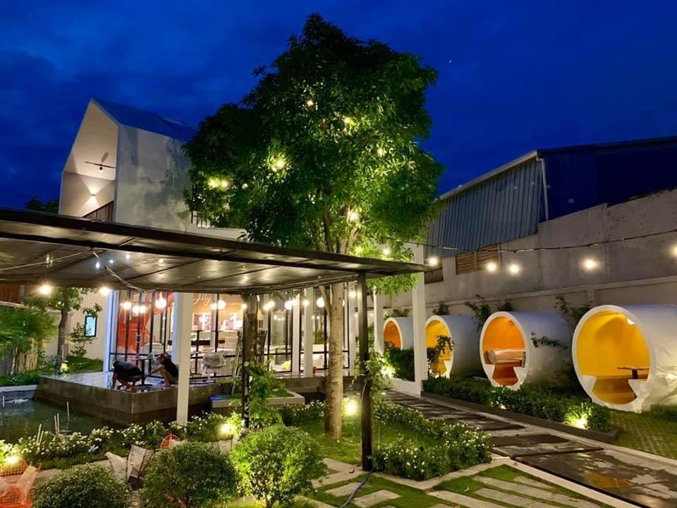 Khu vườn buổi tối với đèn treo, cây xanh, và các gian ngồi hình trụ tròn màu trắng, ánh sáng ấm áp.