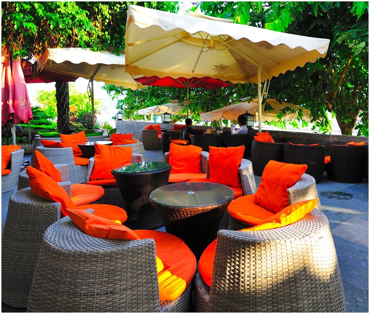 Khu vực ngồi ngo&agrave;i trời với b&agrave;n ghế m&acirc;y v&agrave; đệm cam, c&oacute; &ocirc; che nắng, xung quanh l&agrave; c&acirc;y xanh.