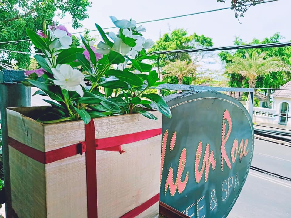 Chậu hoa nhiều m&agrave;u cạnh biển hiệu tr&ecirc;n ban c&ocirc;ng, ph&iacute;a sau l&agrave; c&acirc;y xanh v&agrave; đường phố.