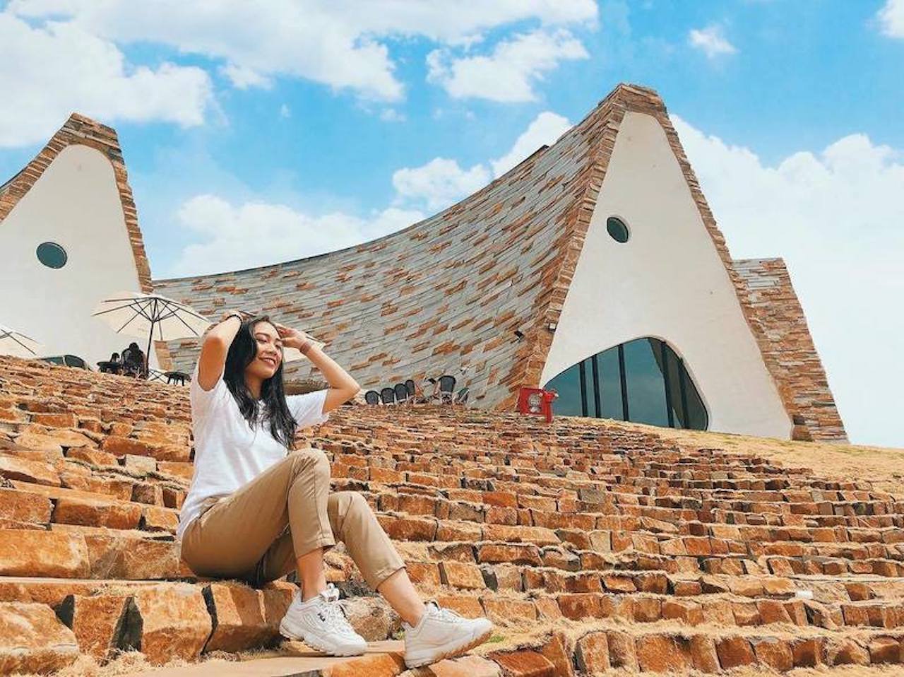 Người ngồi tr&ecirc;n bậc thang đ&aacute; trước t&ograve;a nh&agrave; c&oacute; kiến tr&uacute;c độc đ&aacute;o, dưới bầu trời xanh v&agrave; m&acirc;y trắng.